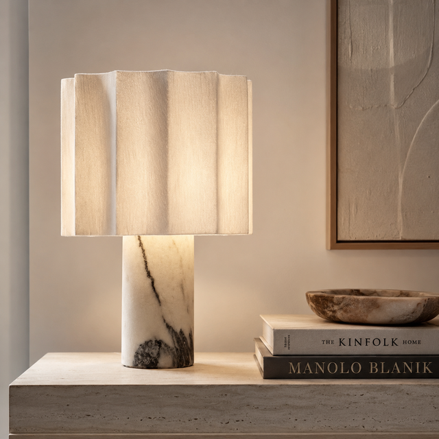 A table lamp with a pleated beige fabric shade and a white marble base with dark veining, styled on a light stone console table next to books and a bowl in a warm interior setting.