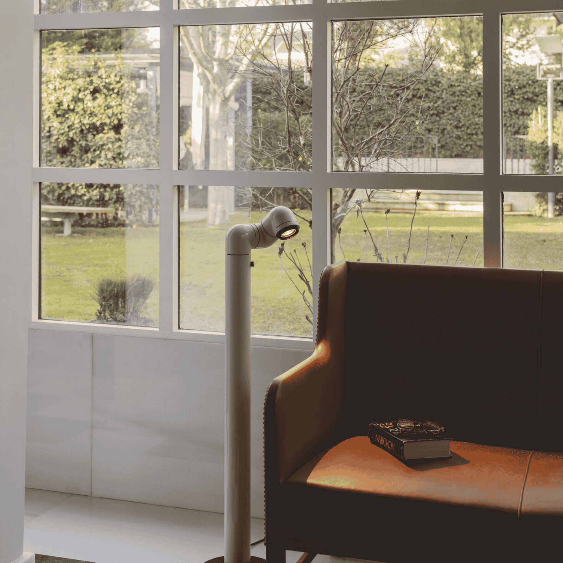 Brown leather chair with a book next to a floor lamp in a room with large windows showing a garden view.