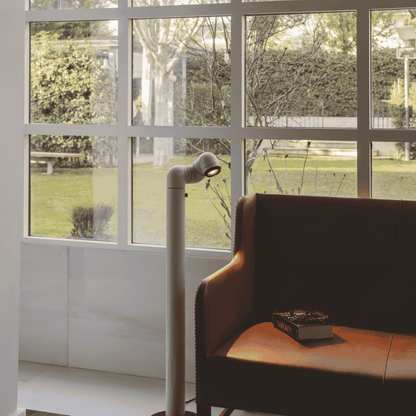 Brown leather chair with a book next to a floor lamp in a room with large windows showing a garden view.