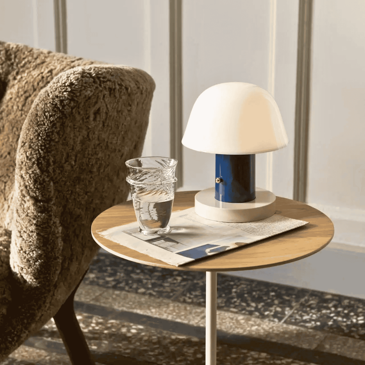 Small round wooden table with a lamp, glass of water, and newspaper next to a textured armchair.