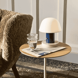 Small round wooden table with a lamp, glass of water, and newspaper next to a textured armchair.