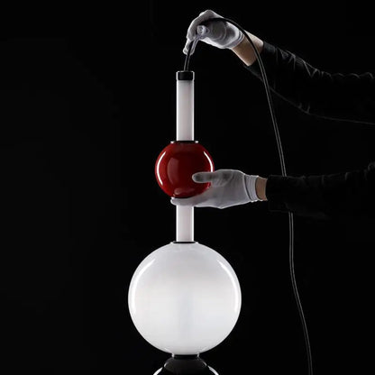 Person in white gloves pouring a red liquid from a small container into a large white sphere on a black background

