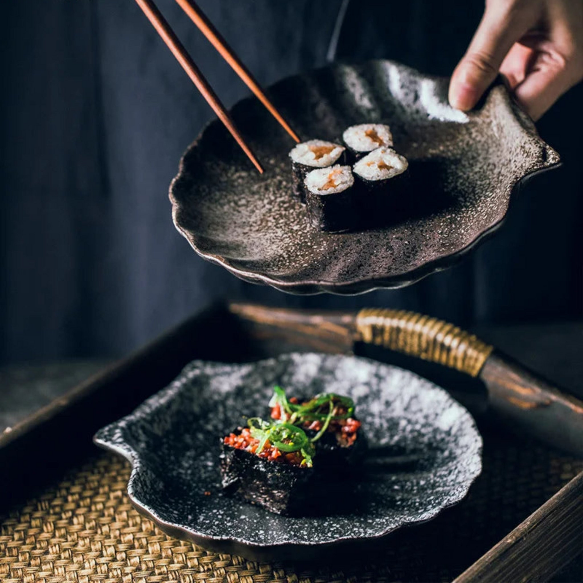 Sushi on a dark textured plate with a hand holding chopsticks, set against a dark background.