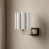 A modern wall-mounted sconce with a fluted white fabric shade and a curved cream-and-brown marble accent, attached to a dark metal bracket on a beige textured wall.