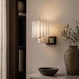 A modern wall sconce with a white pleated shade and a white-and-black marble base, mounted on a beige wall in a styled interior setting.