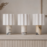 Three table lamps with white pleated drum shades and cylindrical marble bases in cream, beige, and gray tones, arranged on a light stone surface against a soft beige wall.