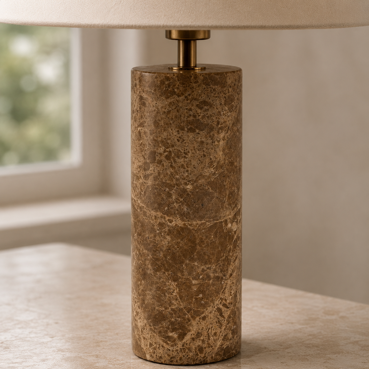 A cylindrical table lamp with a polished brown marble base and a beige fabric drum shade, set on a light stone surface in front of a softly blurred window.