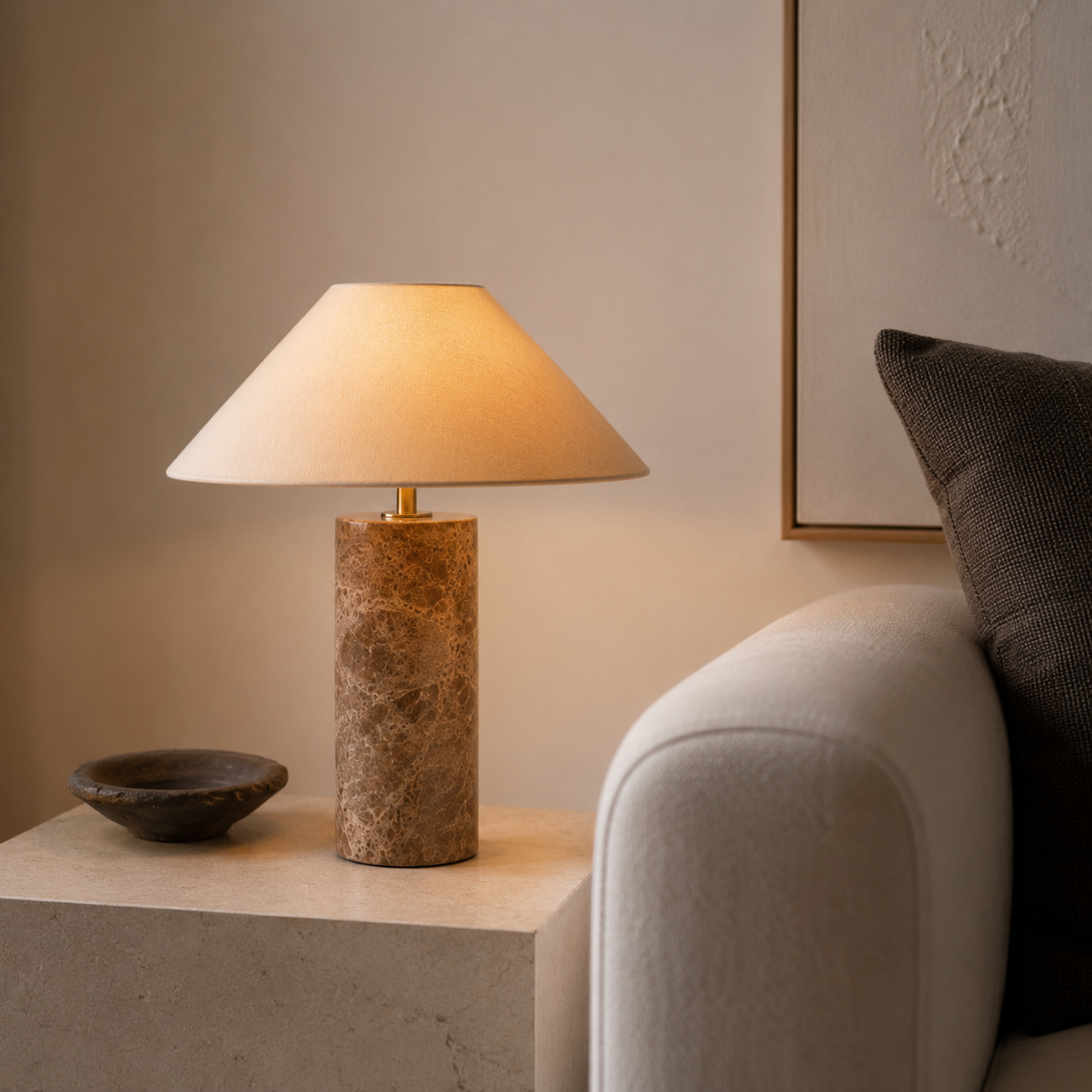 A table lamp with a warm beige fabric drum shade and a cylindrical textured stone base in earthy brown and gold tones, styled on a stone side table next to a sofa in a modern living room setting.