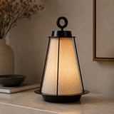A modern table lamp with a black metal frame and a warm beige linen shade, shown lit on a stone surface in a styled interior setting.