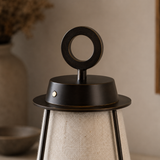 Close-up of a table lamp with a dark bronze metal top featuring a circular ring handle and a textured beige fabric shade, set against a warm neutral background.