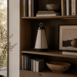 A modern table lamp with a tapered beige fabric shade and black base sits on a wooden bookshelf next to books, bowls, and framed art in a warm, neutral-toned interior.