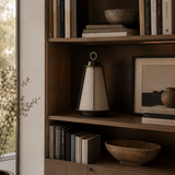 A modern table lamp with a tapered beige fabric shade and black base sits on a wooden bookshelf next to books, bowls, and framed art in a warm, neutral-toned interior.