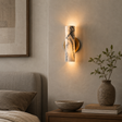 A modern wall sconce with a cylindrical marble-look shade and a gold circular backplate, mounted on a textured beige wall in a bedroom setting next to a bed and a vase with branches.