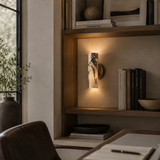 A modern wall sconce with a vertical rectangular white stone or marble column and dark veining, mounted on a warm beige wall and casting a soft glow downward. The sconce is shown in a styled home office setting with wooden shelves, books, and a desk.