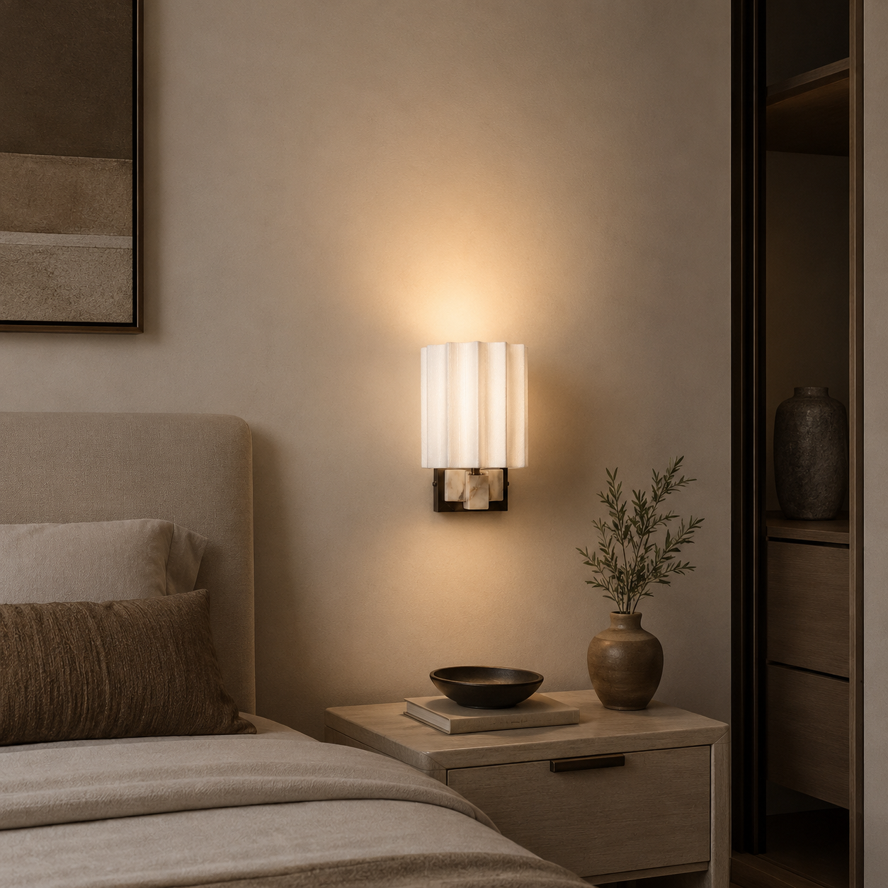 A modern wall-mounted sconce with a white pleated cylindrical shade and a warm glowing light, installed on a beige wall above a wooden nightstand in a bedroom setting.