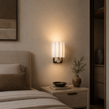 A modern wall-mounted sconce with a white pleated cylindrical shade and a warm glowing light, installed on a beige wall above a wooden nightstand in a bedroom setting.