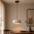 A warm, modern pendant light with a textured stone-look shade and a dark brown cord, hanging over a wooden dining table in a minimalist, warm-toned room.