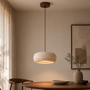A warm, modern pendant light with a textured stone-look shade and a dark brown cord, hanging over a wooden dining table in a minimalist, warm-toned room.