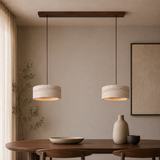 Two modern pendant lights with textured beige drum shades hang over a wooden dining table in a minimalist, warm-toned room with a beige wall and neutral decor.