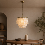A tiered chandelier pendant light with a warm brass frame and layered translucent glass discs, hanging over a wooden dining table in a softly lit, neutral-toned room.