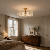 A modern semi-flush mount ceiling light fixture with a gold brass central canopy and textured, petal-like clear glass shades, shown installed in a warm, modern bedroom setting.