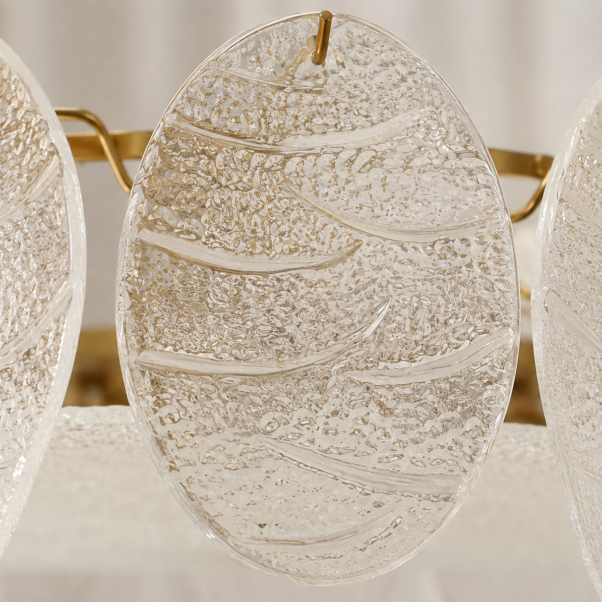 A close-up of a clear textured glass wall sconce with organic wavy patterns and a gold hanging loop, shown against a soft neutral background.