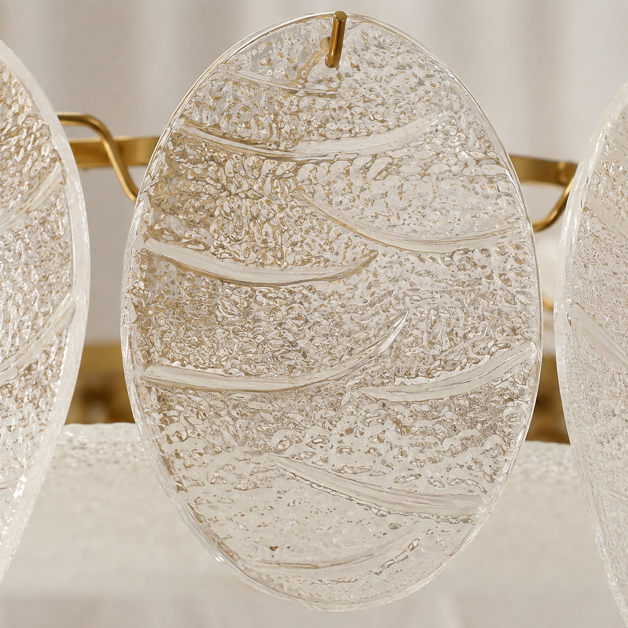 A close-up of a clear textured glass wall sconce with organic wavy patterns and a gold hanging loop, shown against a soft neutral background.