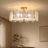 A modern semi-flush mount ceiling light fixture with a gold-finished ceiling mount and frame, featuring multiple textured, petal-like clear glass shades that glow with warm light. The fixture is shown installed on a beige ceiling in a styled room with curtains, a console table, and a framed artwork.