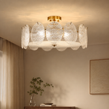 A modern semi-flush mount ceiling light fixture with a gold-finished ceiling mount and frame, featuring multiple textured, petal-like clear glass shades that glow with warm light. The fixture is shown installed on a beige ceiling in a styled room with curtains, a console table, and a framed artwork.