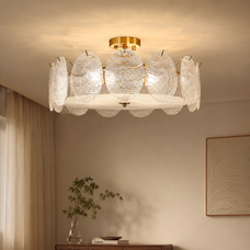 A modern semi-flush mount ceiling light fixture with a gold-finished ceiling mount and frame, featuring multiple textured, petal-like clear glass shades that glow with warm light. The fixture is shown installed on a beige ceiling in a styled room with curtains, a console table, and a framed artwork.