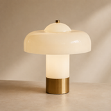 A mushroom-shaped table lamp with a frosted white glass shade, a glowing cylindrical body, and a brushed brass base, set against a warm beige background.