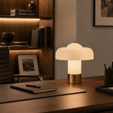 A mushroom-shaped table lamp with a glowing white glass shade and a brushed gold metal base, placed on a dark wooden desk in a modern office setting.