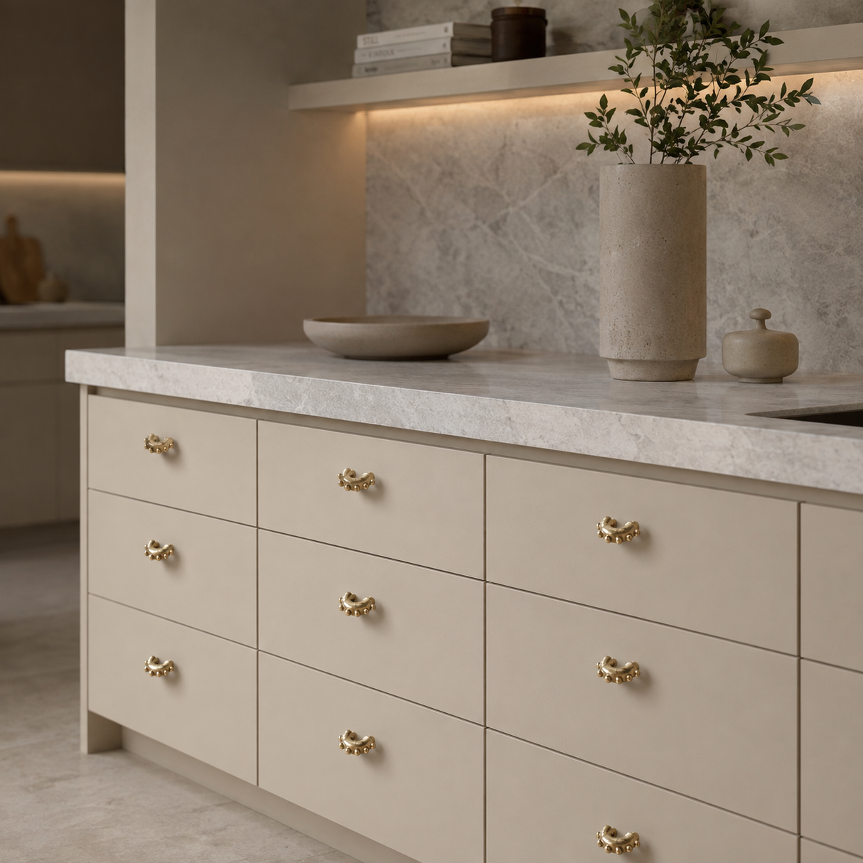 A modern kitchen cabinet with nine cream-colored drawers featuring decorative gold handles, set under a light gray marble countertop with a matching stone backsplash.