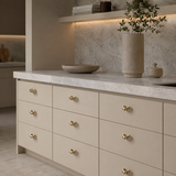 A modern kitchen cabinet with nine cream-colored drawers featuring decorative gold handles, set under a light gray marble countertop with a matching stone backsplash.