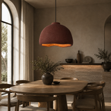 A rustic terracotta-colored dome pendant light hangs over a wooden dining table in a warm, modern dining room with neutral walls and natural light from an arched window.