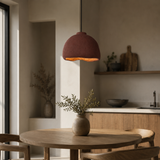 A terracotta-colored dome pendant light hangs over a round wooden dining table in a warm, modern kitchen setting.