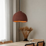 A dome-shaped pendant light with a textured rust-brown shade and a warm glowing orange interior, hanging over a dining table in a modern, neutral-toned room.
