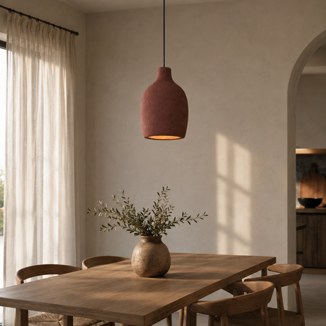 A terracotta-colored pendant light hangs over a wooden dining table in a warm, modern dining room with cream walls, sheer curtains, and a vase of greenery on the table.