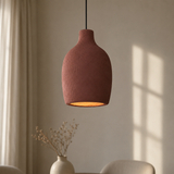 A textured terracotta pendant light hanging in a softly lit room with a beige wall, window shadows, and a small vase of dried stems below.