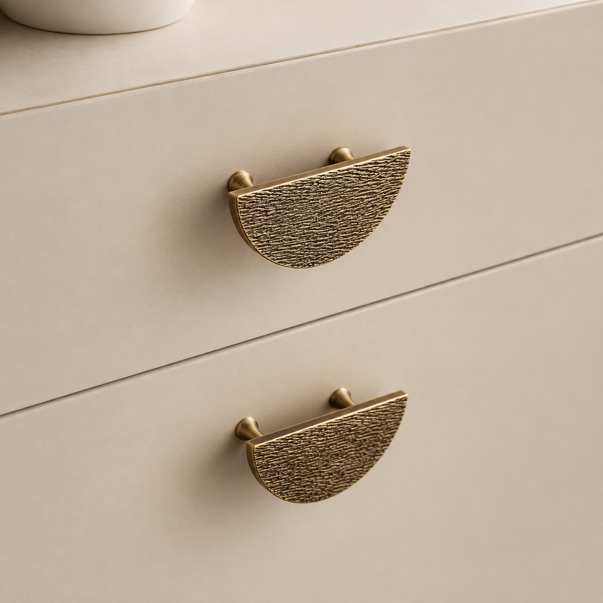 Close-up of two textured brass semi-circular drawer pulls on a light beige wooden cabinet.