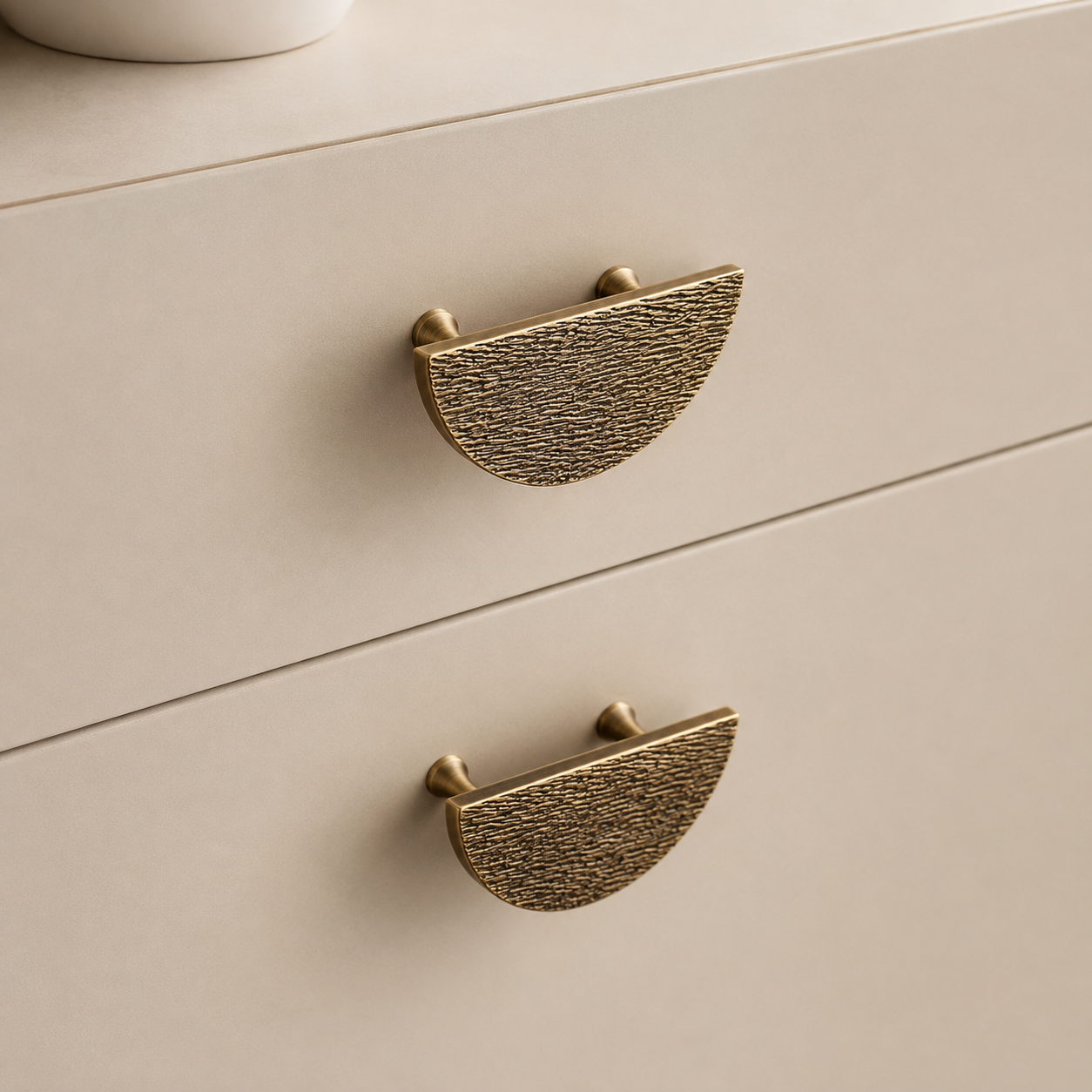 Close-up of two textured brass semi-circular drawer pulls on a light beige wooden cabinet.