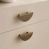 Close-up of two textured brass semi-circular drawer pulls on a light beige wooden cabinet.