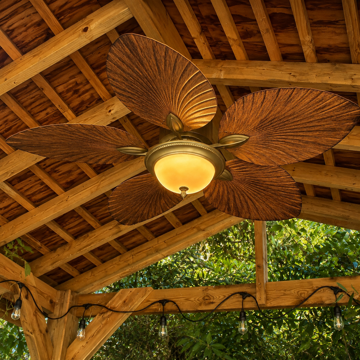 A five-blade ceiling fan with textured, fan-shaped brown blades and a warm glowing dome light fixture, mounted on a wooden porch ceiling with string lights and green foliage visible below.