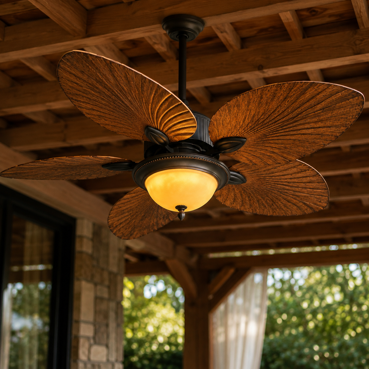 A decorative ceiling fan with five textured, fan-shaped blades in a warm brown finish, suspended under a wooden porch ceiling with a glowing warm white globe light.