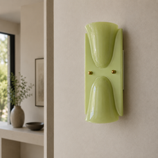 A modern wall-mounted sconce with a glossy light green glass shade and two small gold-toned side knobs, shown mounted on a beige wall in a minimalist interior setting.