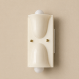 A modern wall-mounted light fixture with a cream-colored sculptural frame, two small gold screws, and white globe bulbs on either side, set against a solid beige background.