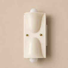 A modern wall-mounted light fixture with a cream-colored sculptural frame, two small gold screws, and white globe bulbs on either side, set against a solid beige background.
