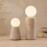 Two modern table lamps with textured stone bases and glowing frosted spherical shades, shown on a warm beige surface against a softly textured neutral background.