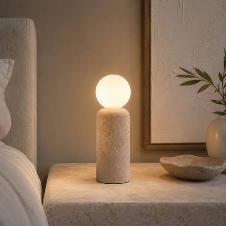 A modern stone table lamp with a cylindrical textured base and a glowing spherical shade, placed on a matching stone bedside table in a softly lit bedroom setting.