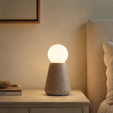 A modern table lamp with a textured concrete cone base and a glowing spherical shade, placed on a bedside table next to a bed in a warm, neutral-toned bedroom.